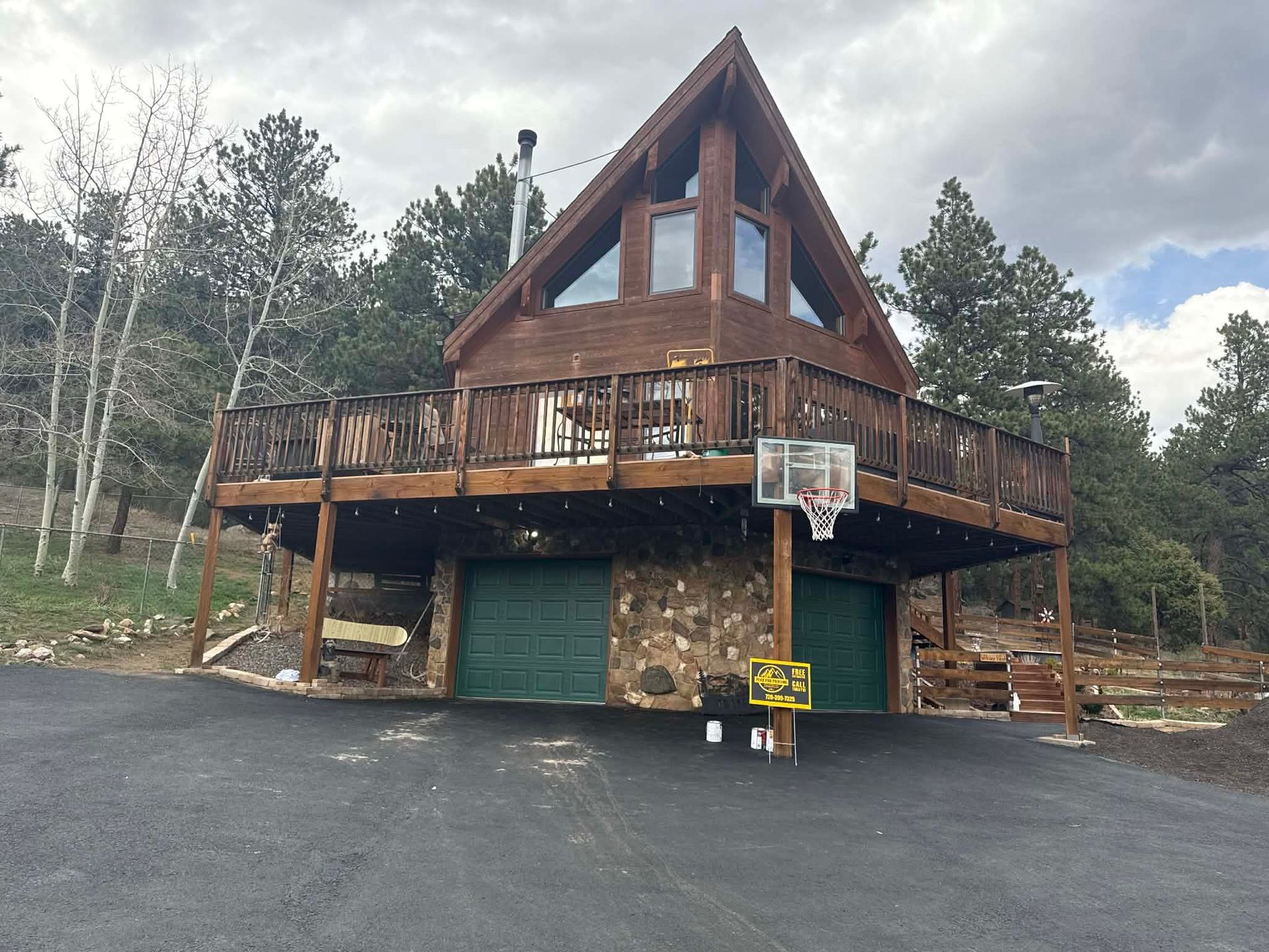 A large A-frame cabin with stone base, deck, and basketball hoop in a wooded driveway.