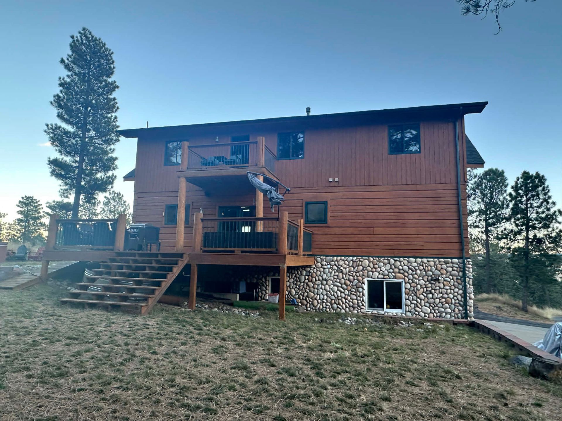 Wood-sided house with a front deck and stairs, set on a grassy hill at dusk.