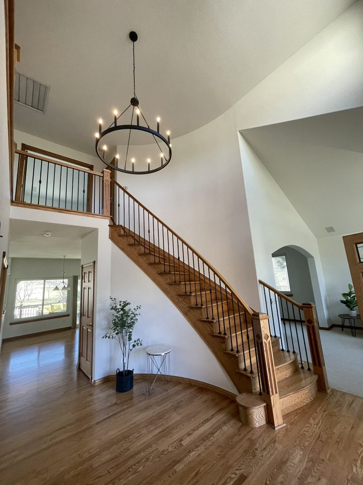 A grand foyer with light wood floors, a wooden staircase with black railings, and a large circular chandelier overhead.