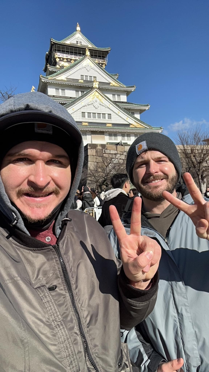 Two smiling men in beanies and jackets pose for a selfie, giving peace signs in front of Osaka Castle on a sunny day.