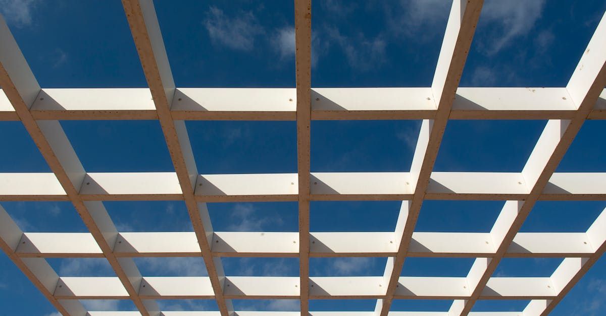 A white wooden pergola frame against a blue sky with soft, wispy clouds.