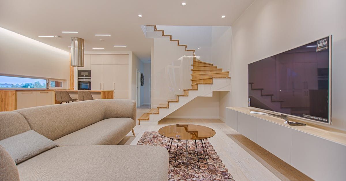 A modern living room with a neutral sectional sofa, a floating TV console, a wooden staircase, and an open kitchen.