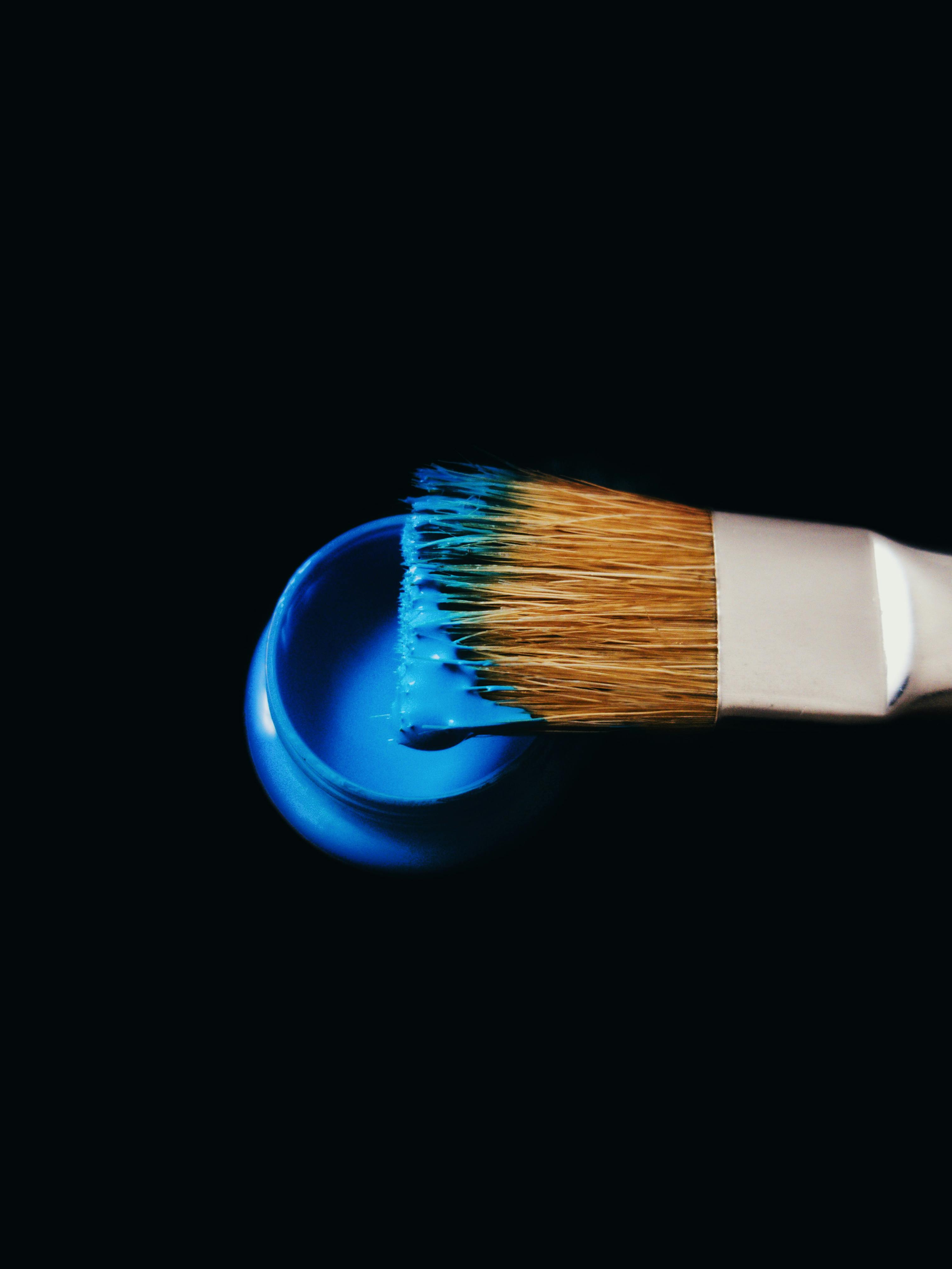A flat-head paintbrush with blue paint on its bristles hovering over a small, matching blue paint container on black.