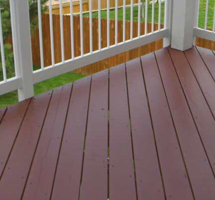 A close-up view of a dark brown painted wooden deck with white railings, set against a blurred green yard and wood fence.
