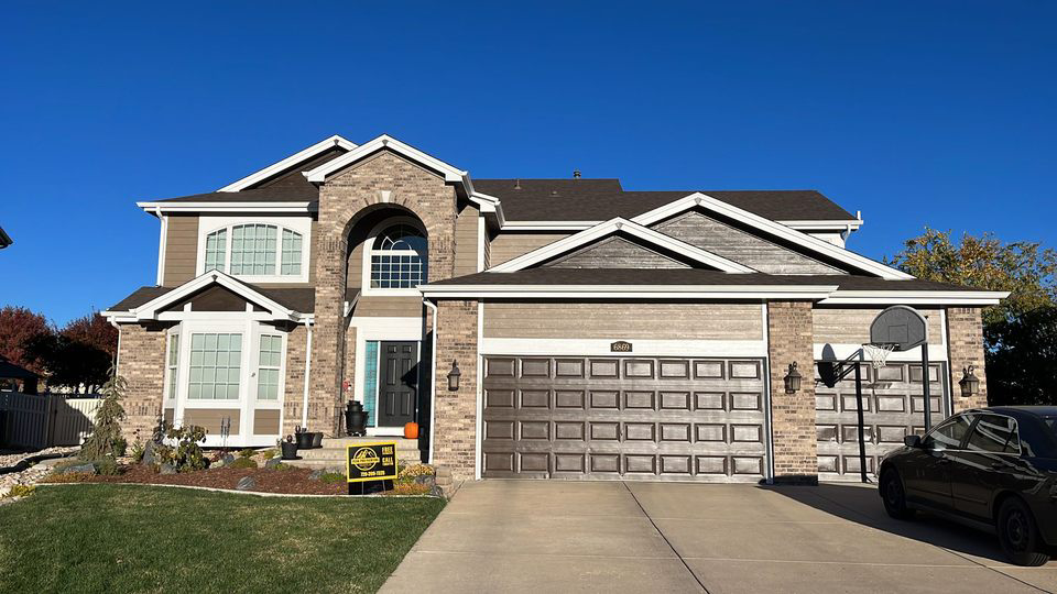 Suburban two-story brick house with a three-car garage under a clear blue sky.