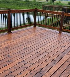 A stained, wooden deck with black metal railings overlooking a pond and green lawn.