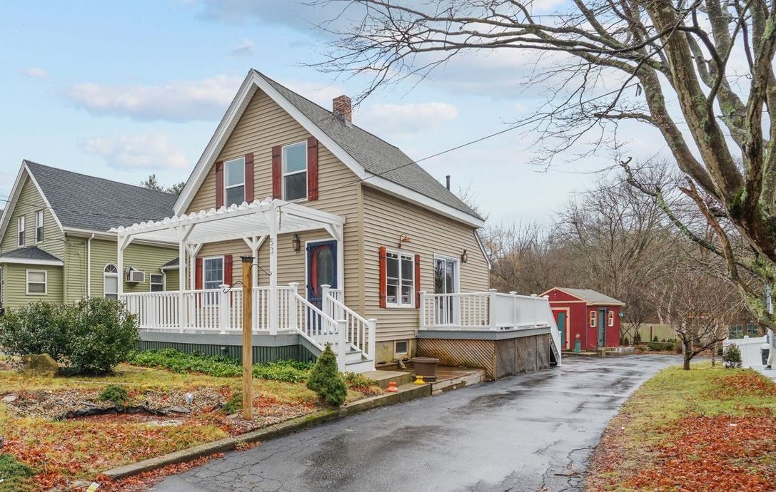 Tan house with white porch and red shutters, next to a green house. Driveway curves to a red shed.
