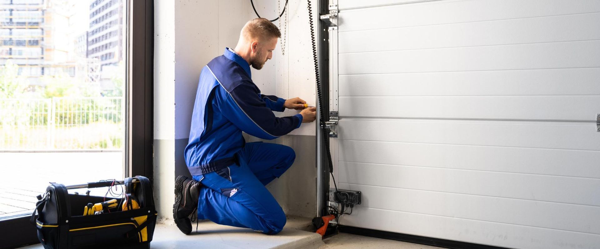 A Man Is Kneeling Down In A Garage Fixing A Garage Door — Mick’s Garage Doors In Alice River, QLD