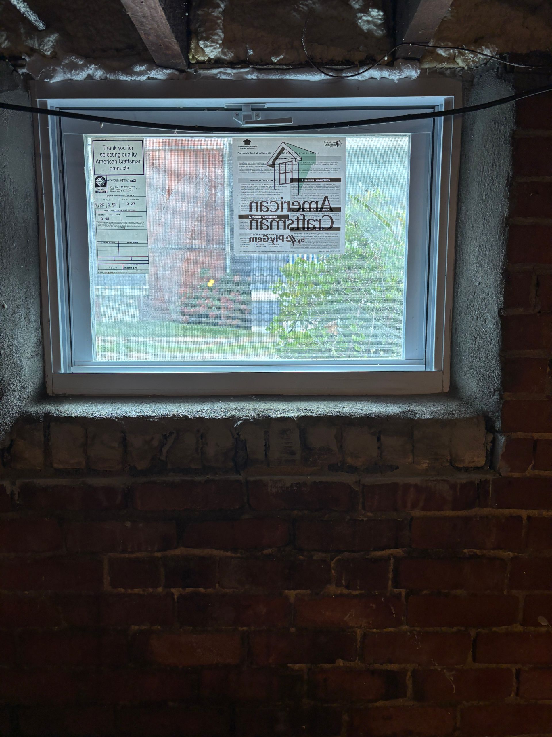 A white basement window set into a brick wall, with a sticker from an American construction company on the glass.