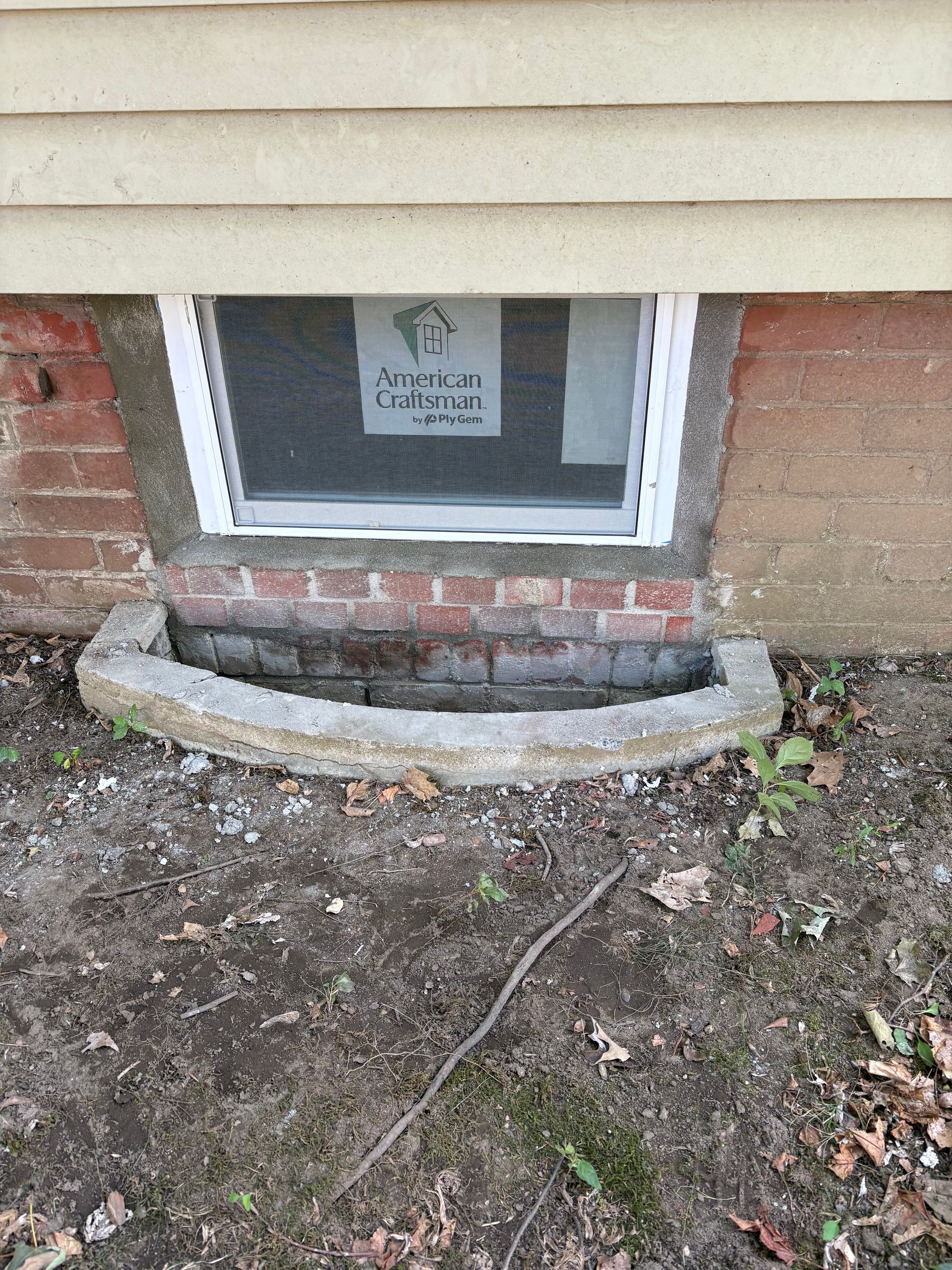 A white basement window with a screen and a concrete window well set into a brick and siding exterior wall.
