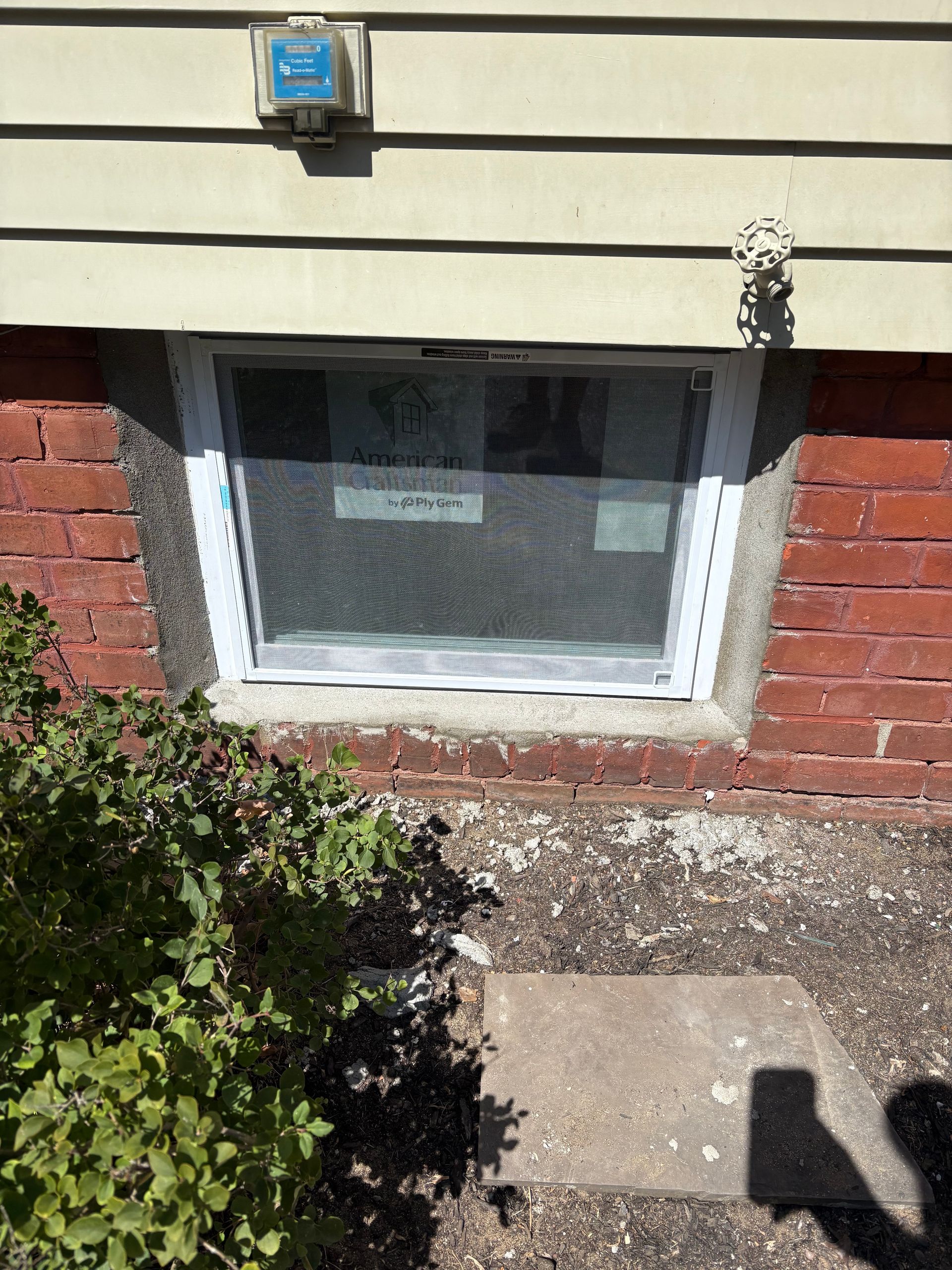 A basement window with a screen is set into a red brick foundation below horizontal beige siding.