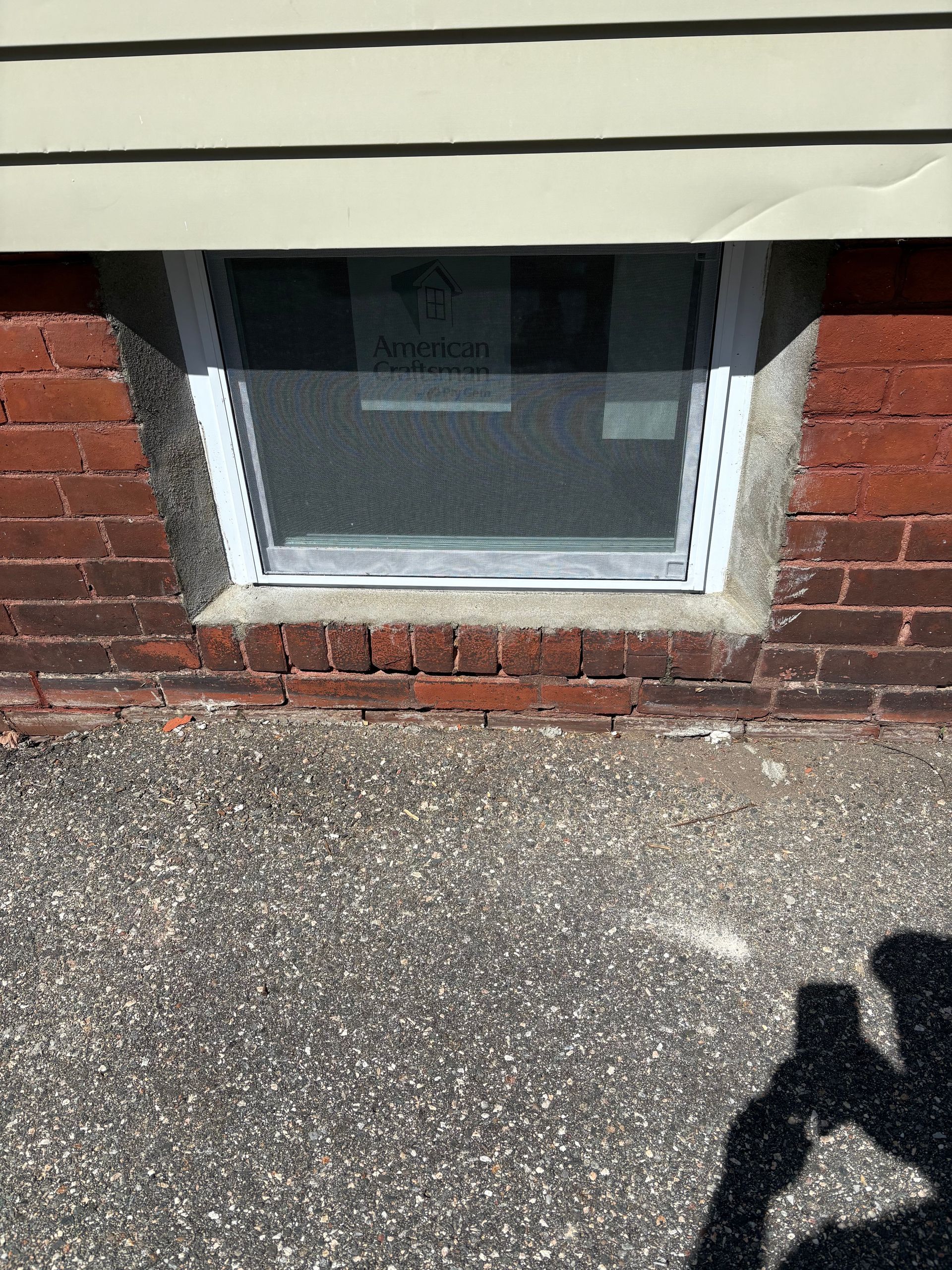 A rectangular basement window with a white frame and mesh screen, set into a red brick foundation below beige siding.