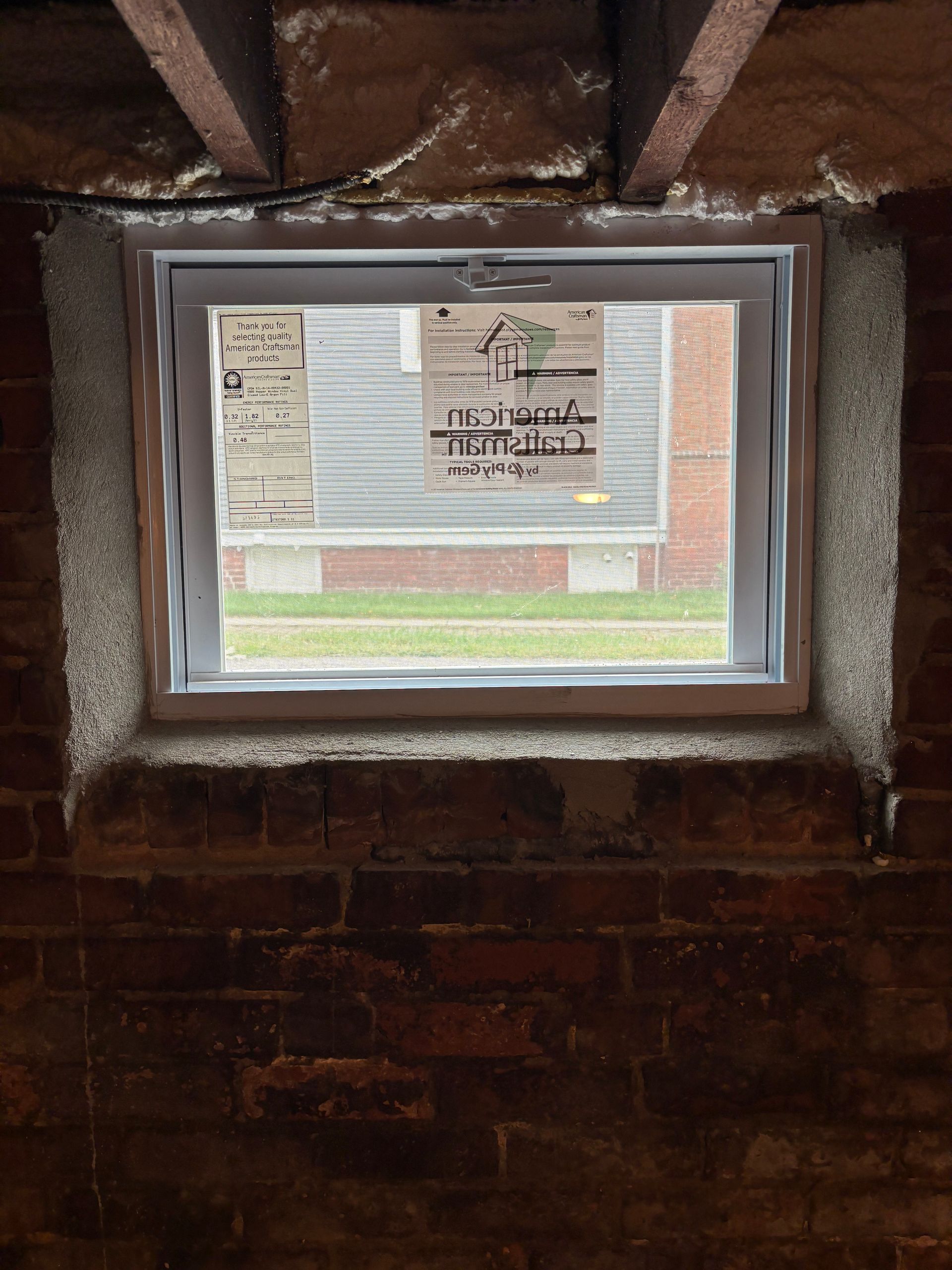 A new white vinyl basement window installed in a brick foundation wall, with a manufacturer sticker on the glass.