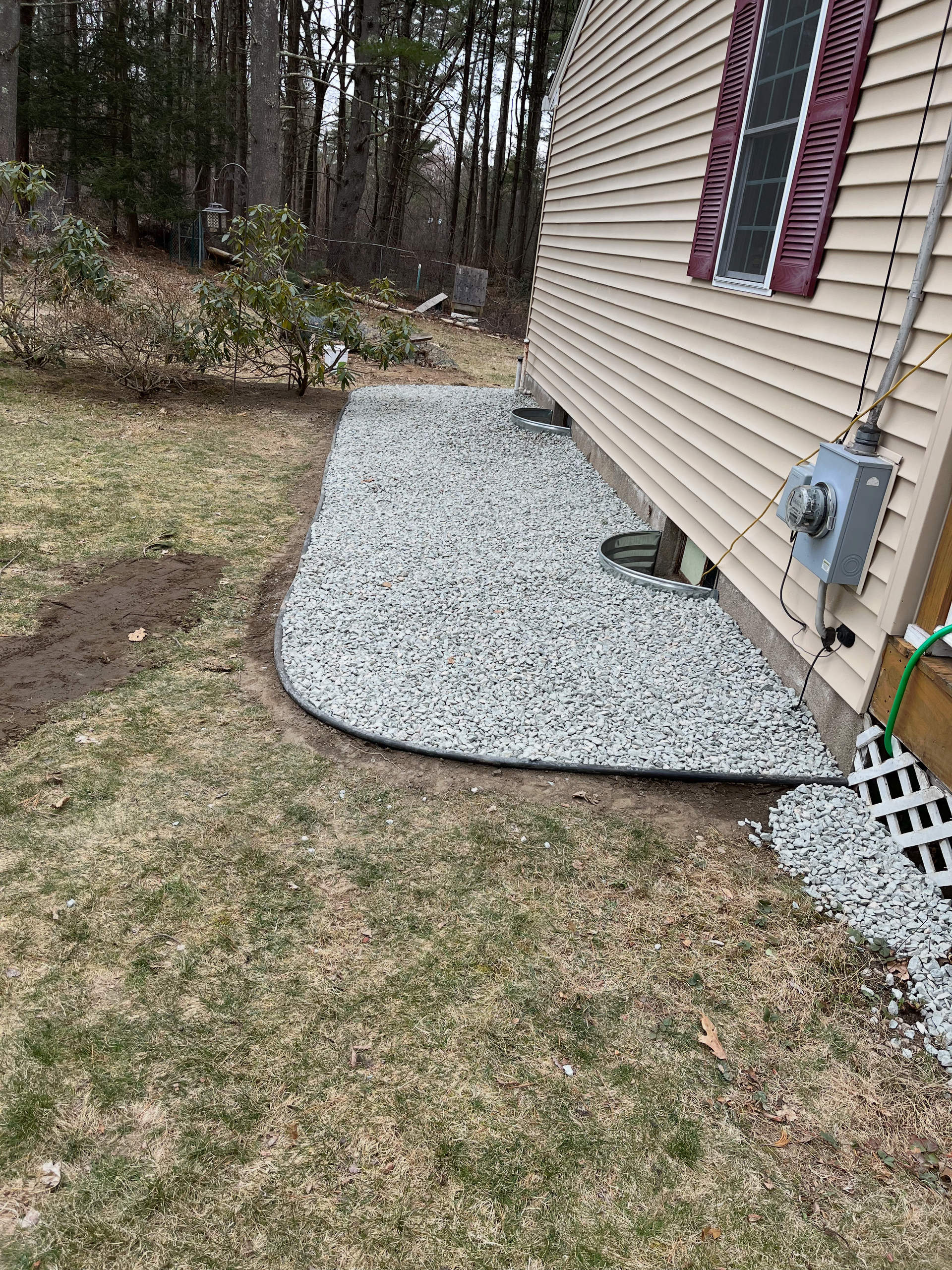 A gravel landscaping border runs along the side of a beige house, curving into the lawn near a white lattice structure.