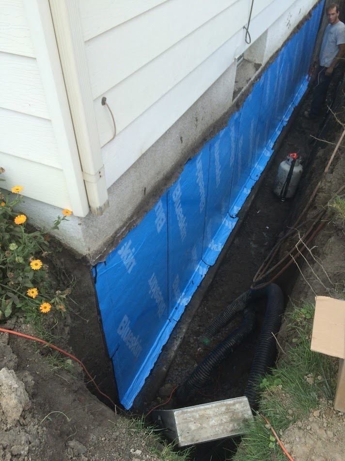 A foundation wall with blue waterproof membrane being installed in a trench next to a house exterior.