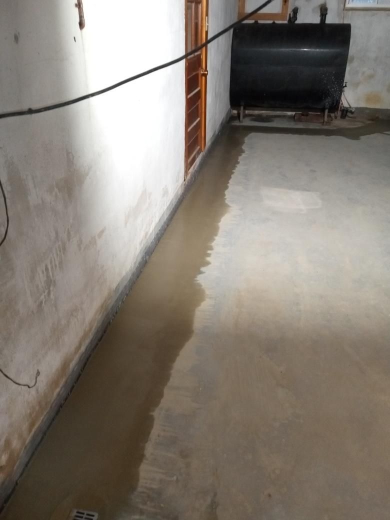 A flooded concrete basement floor with water pooling along the wall near a doorway and a dark storage tank.