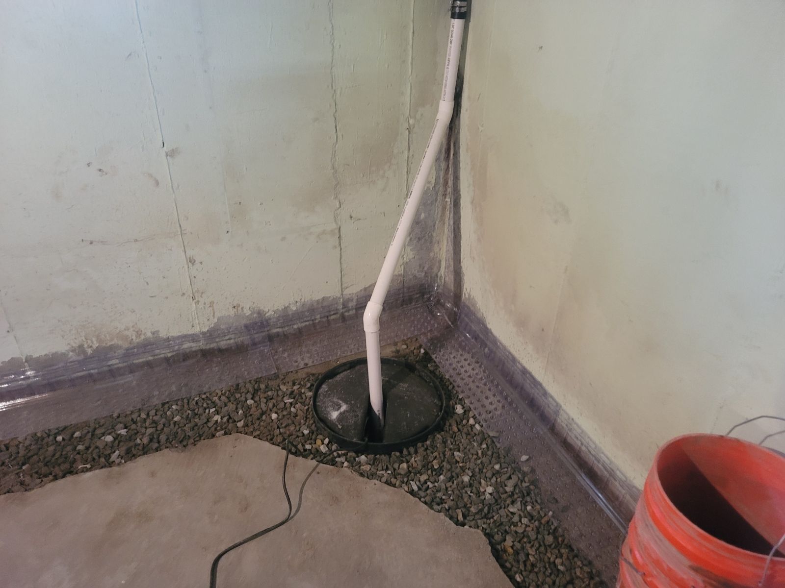 A white PVC pipe rises from a sump pump basin in a gravel-filled corner of a basement with pale walls.
