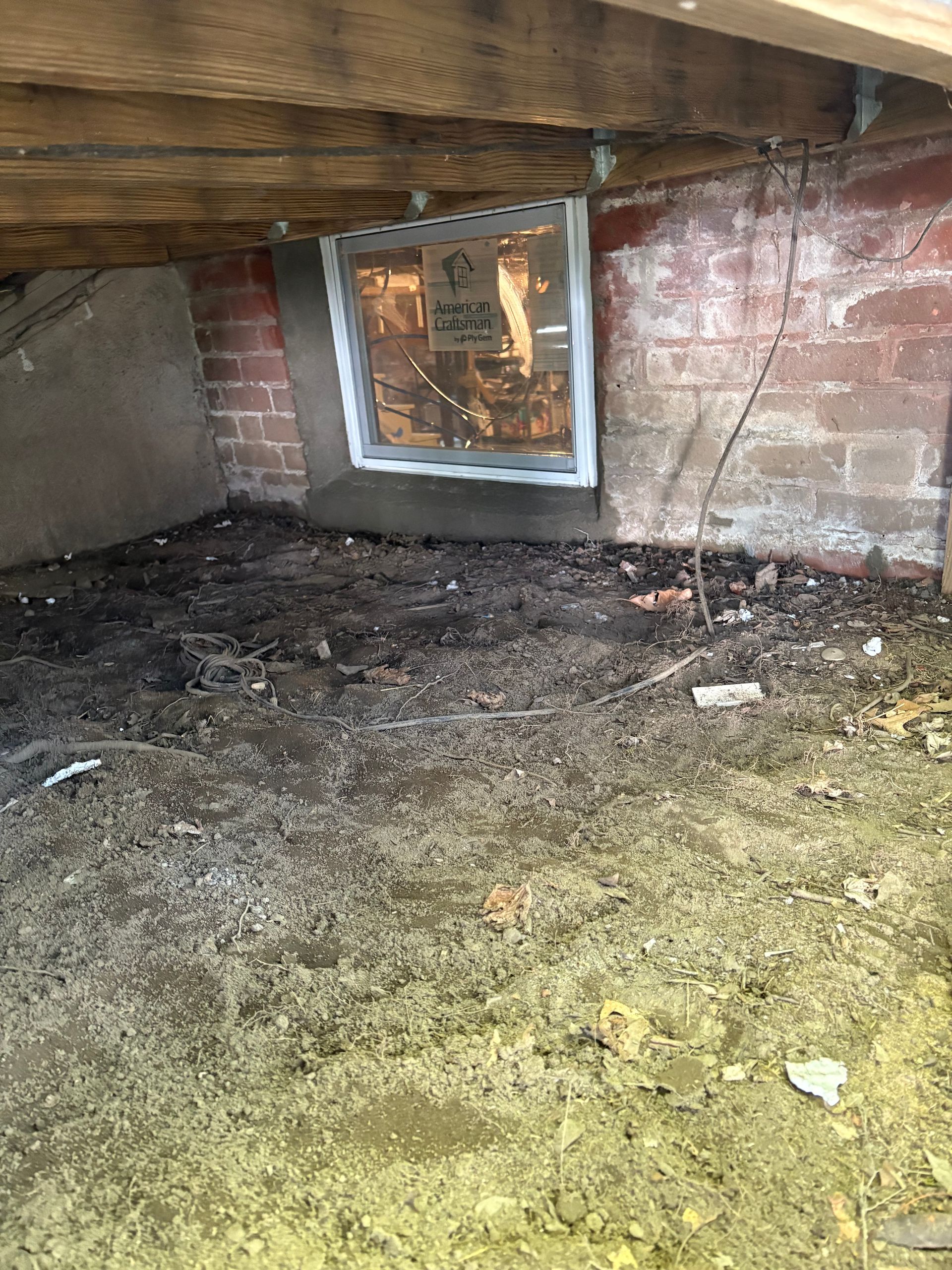 A dirt-floored crawlspace with a white-framed window set into a brick foundation wall beneath wooden floor joists.