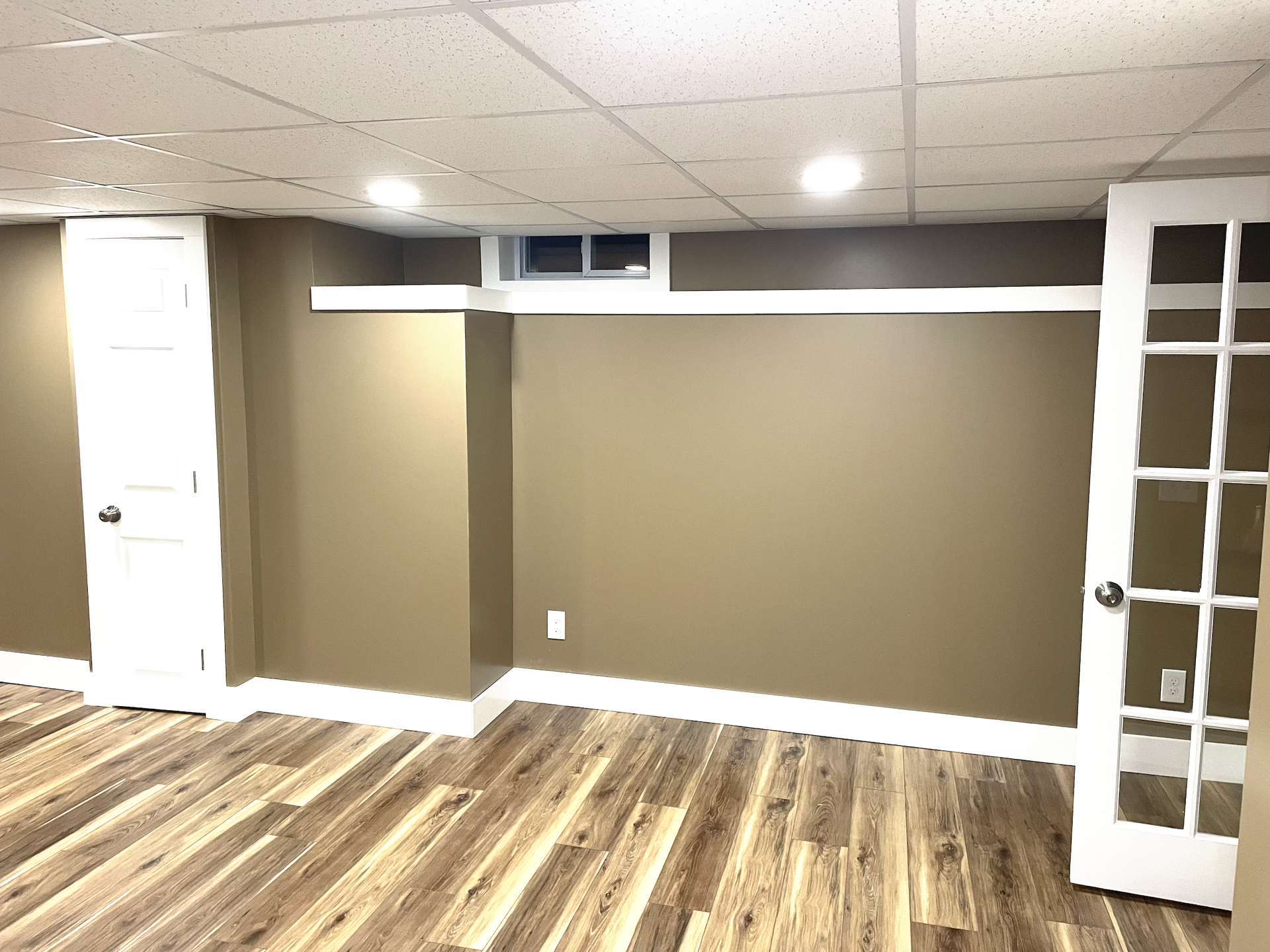 A basement room with tan walls, white trim, a drop ceiling, wood-look flooring, a small window, and two white doors.