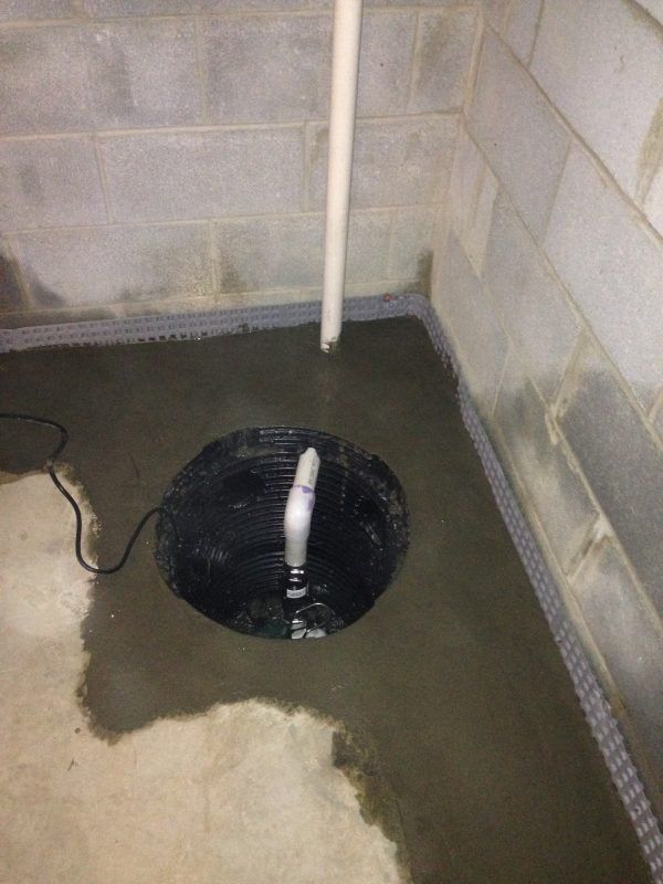 Sump pump basin installed in a concrete basement floor corner with drainage piping along the wall base.