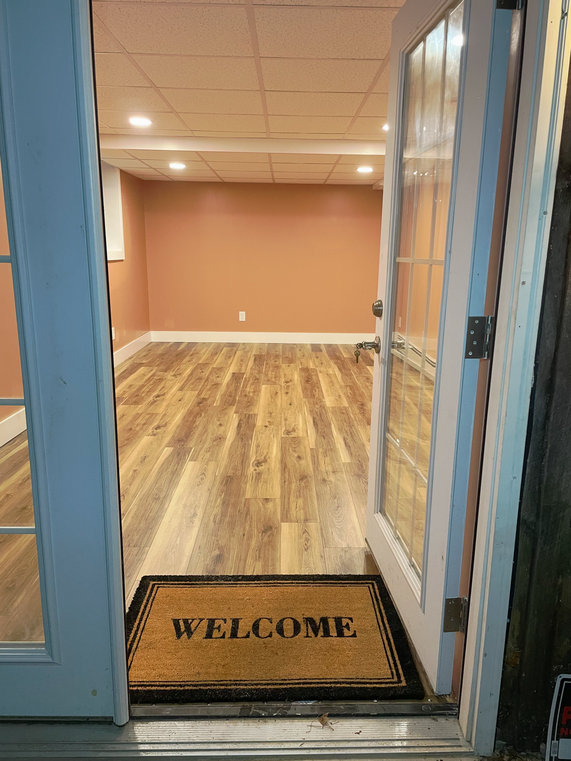Open French doors lead into a basement room with light-colored wood flooring, peach-colored walls, and a welcome mat.