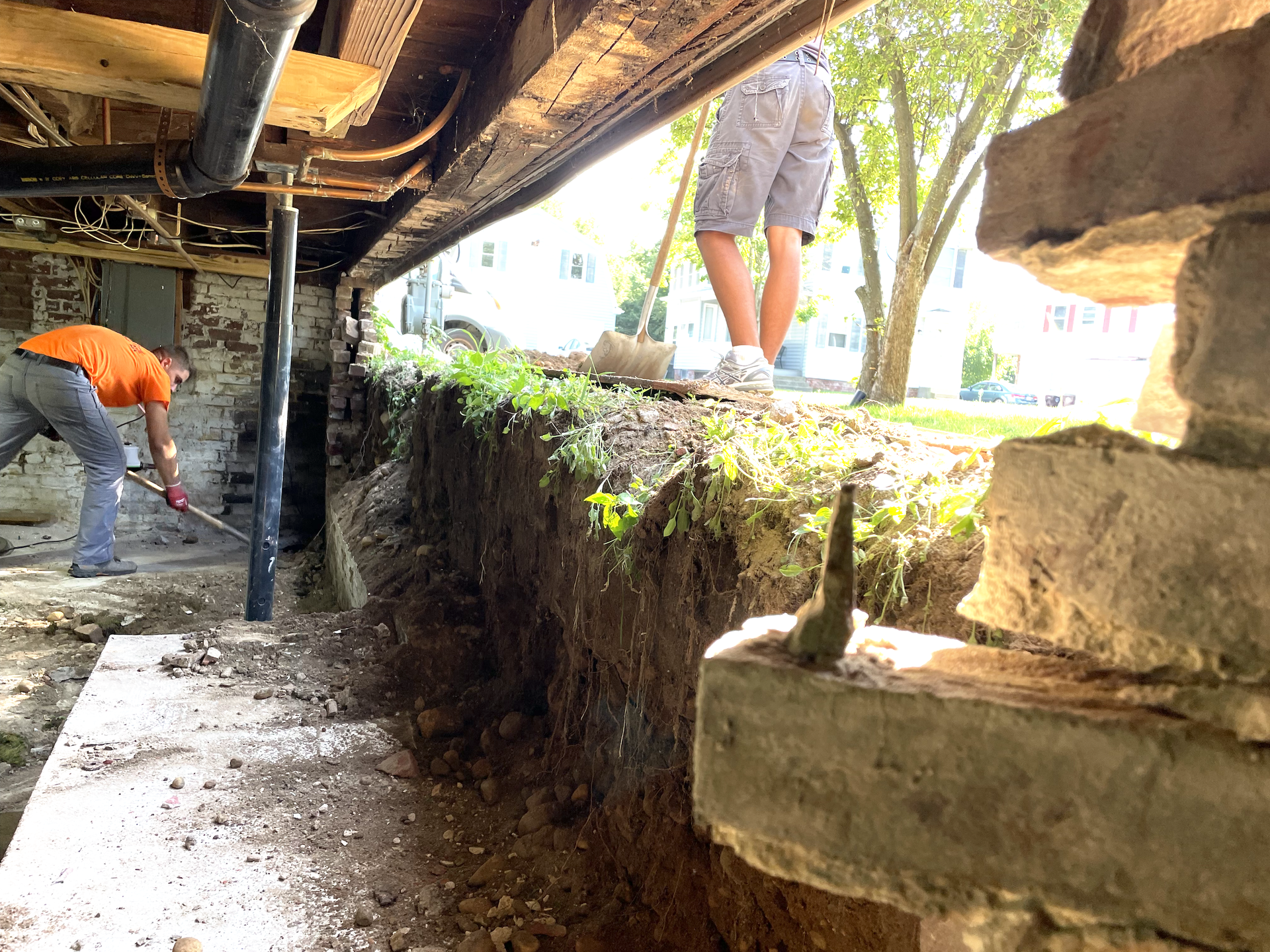 Two people work in a dirt crawl space beneath a building, excavating along a stone foundation wall.