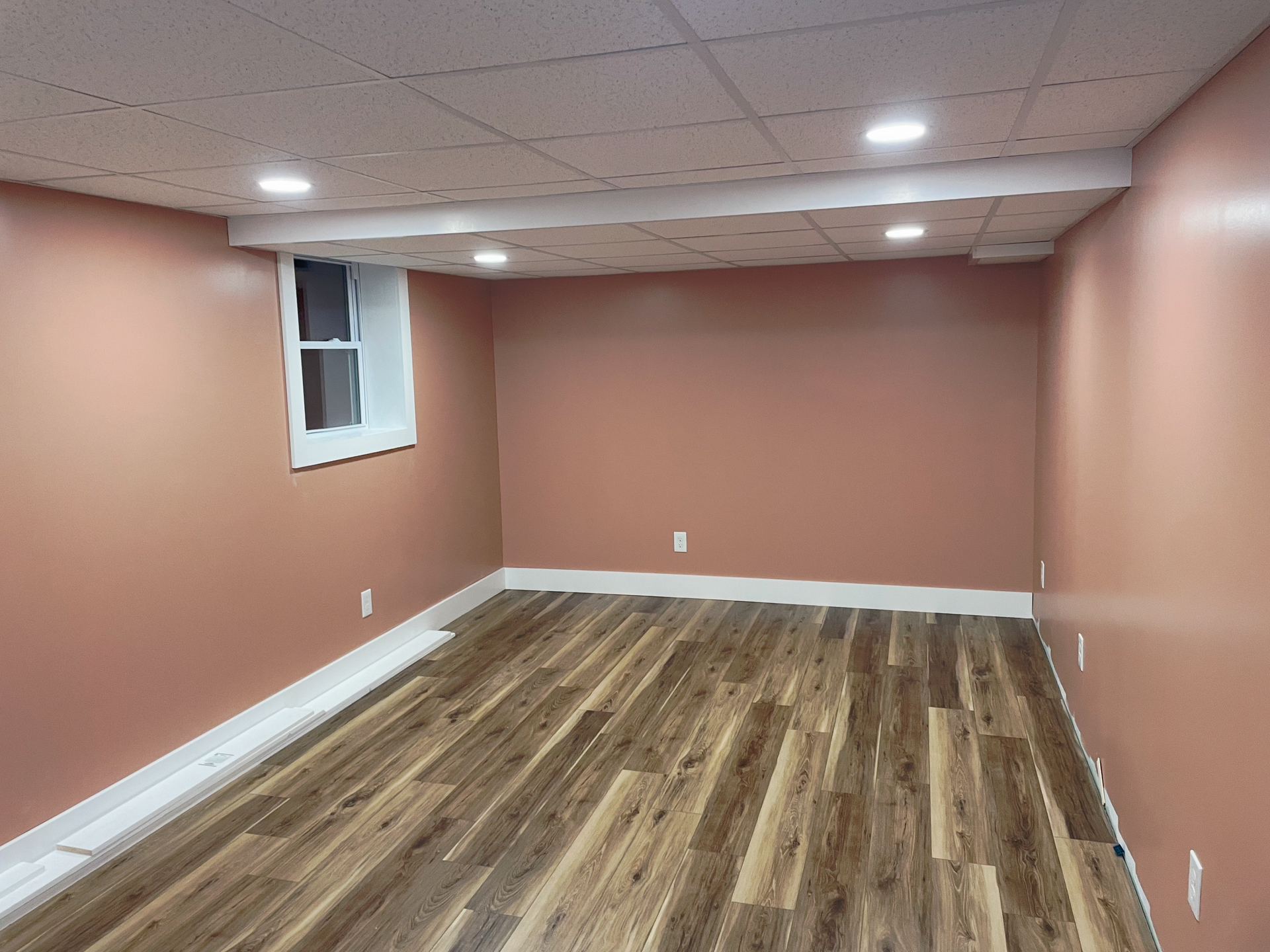 An empty basement room with wood-patterned flooring, dusty rose walls, a small window, and a white drop ceiling.
