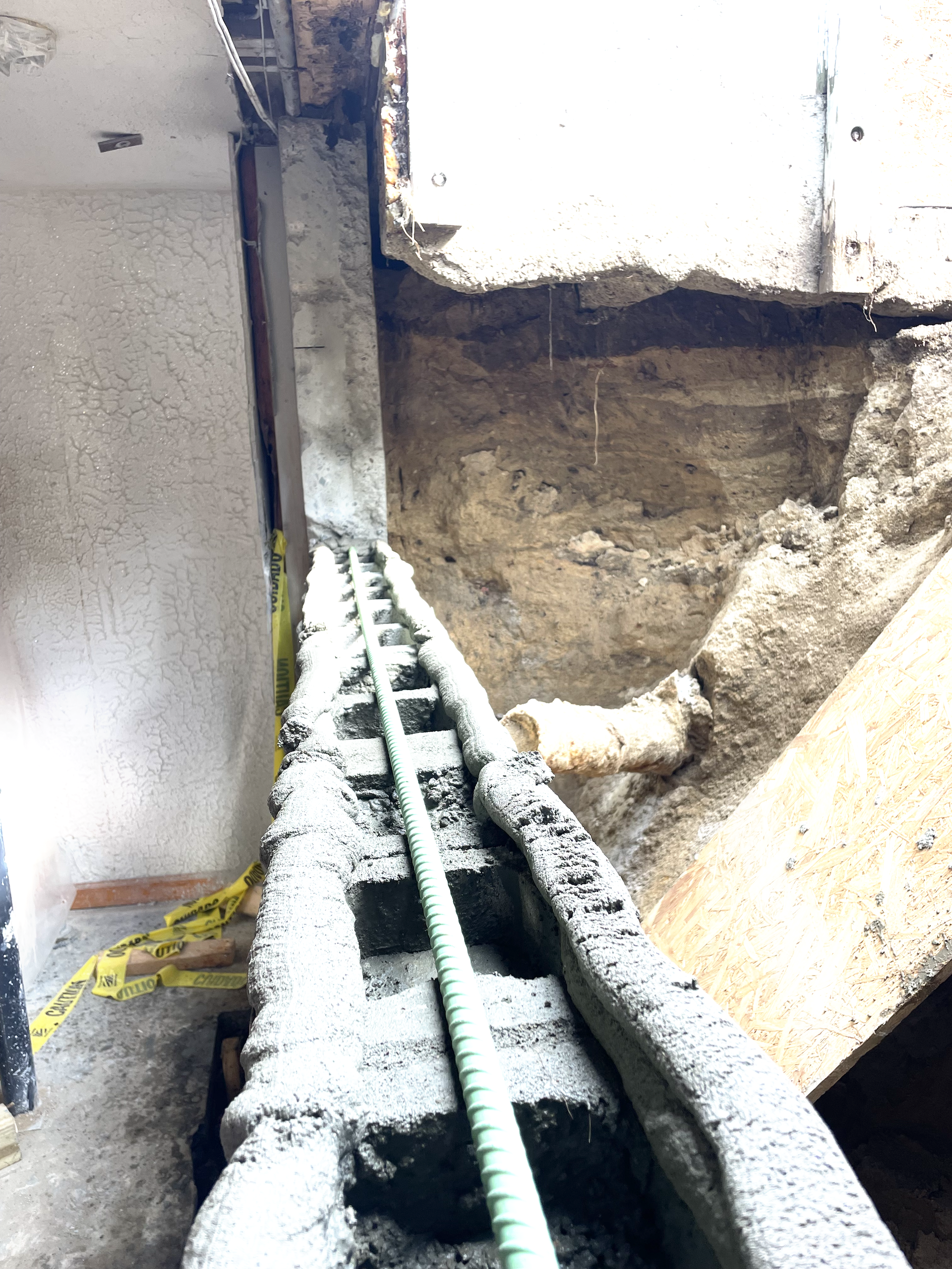 A partially constructed concrete block wall sits in a trench against an excavated dirt wall, reinforced with green rebar.
