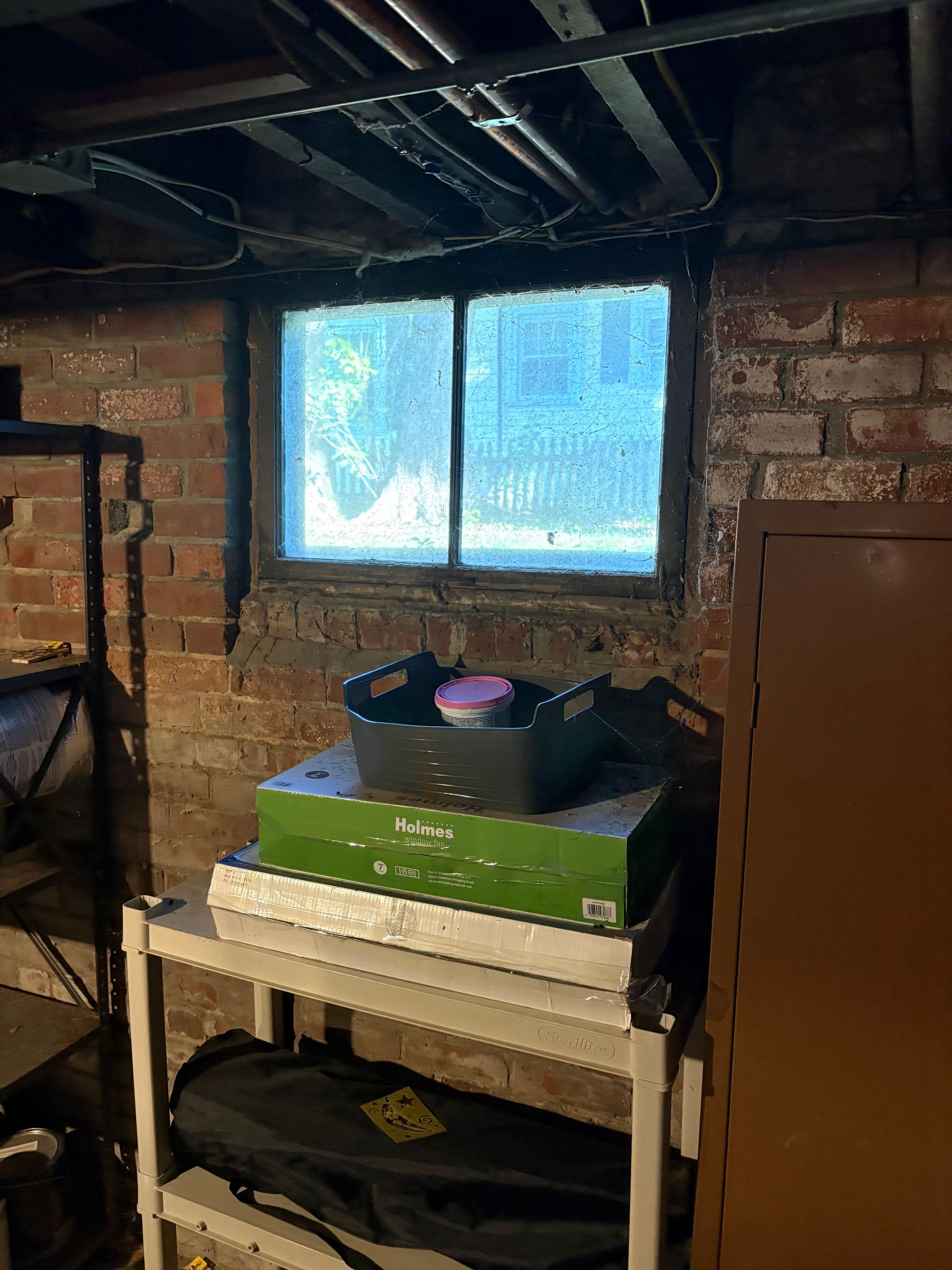 A small, rectangular window set into a brick basement wall above a white utility shelf holding stacked items and a bin.