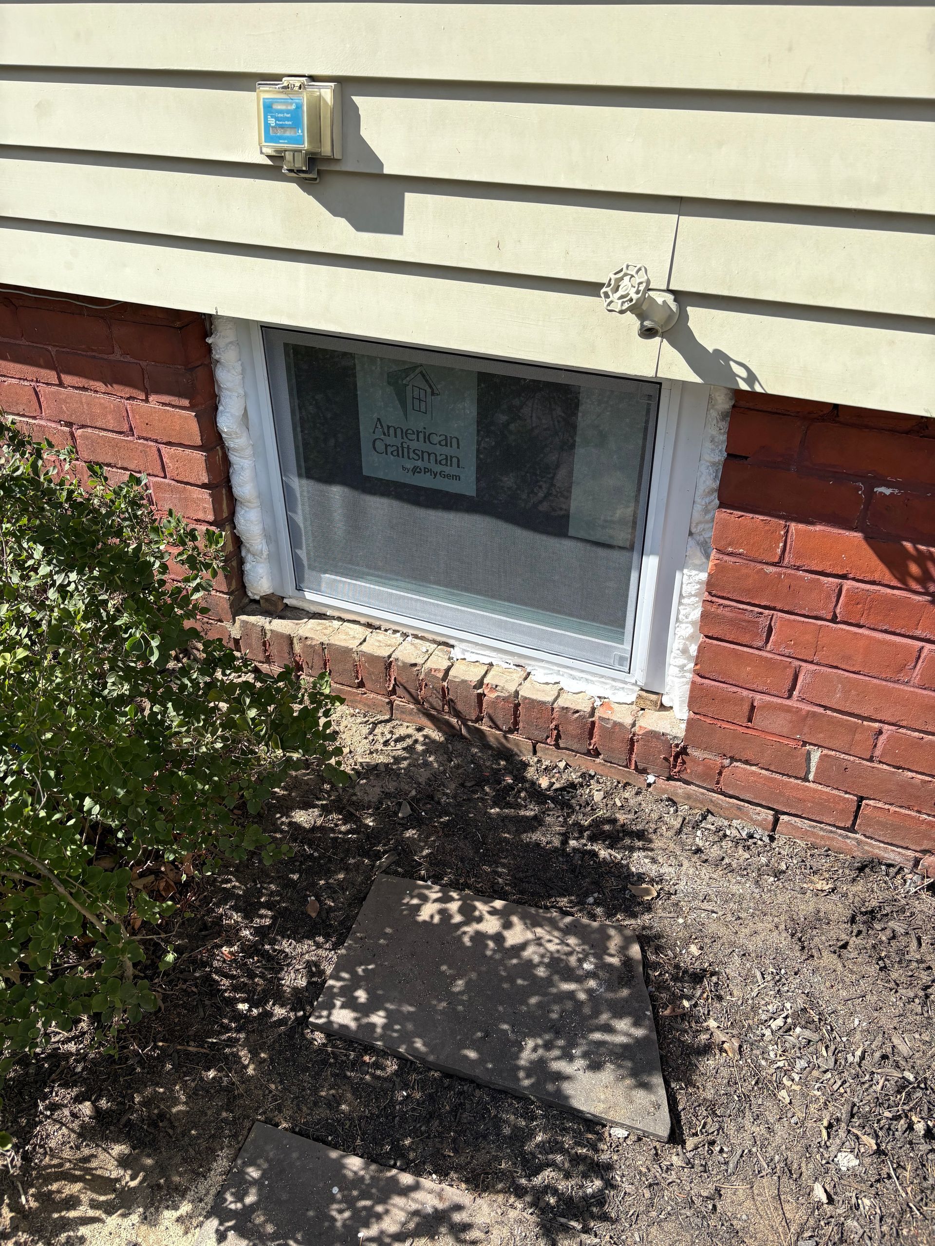 A white-framed basement window set into a red brick foundation under beige siding on the exterior of a house.