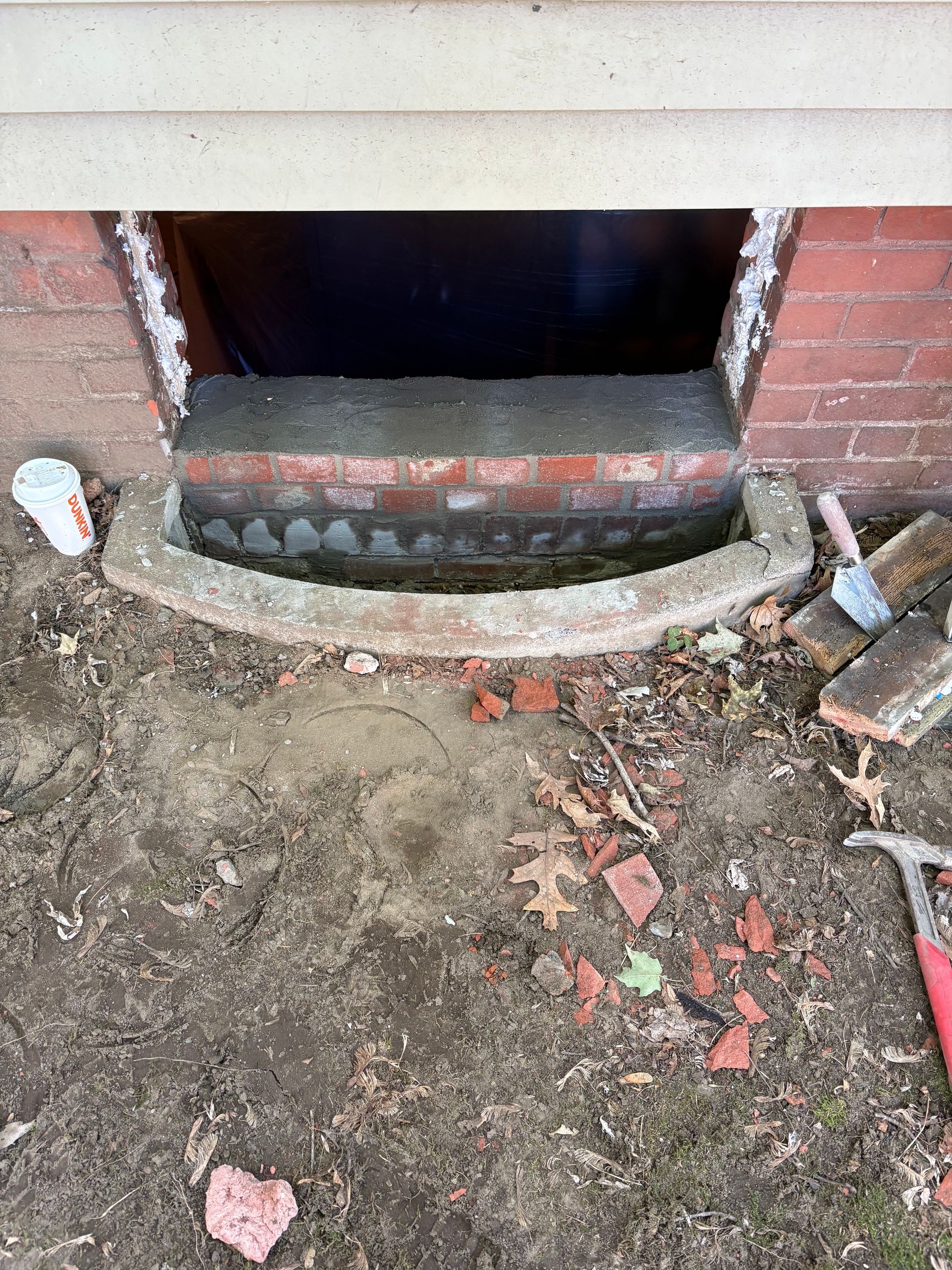 A newly constructed brick curb sits in front of a basement window well, with a trowel and masonry tools nearby on the dirt.