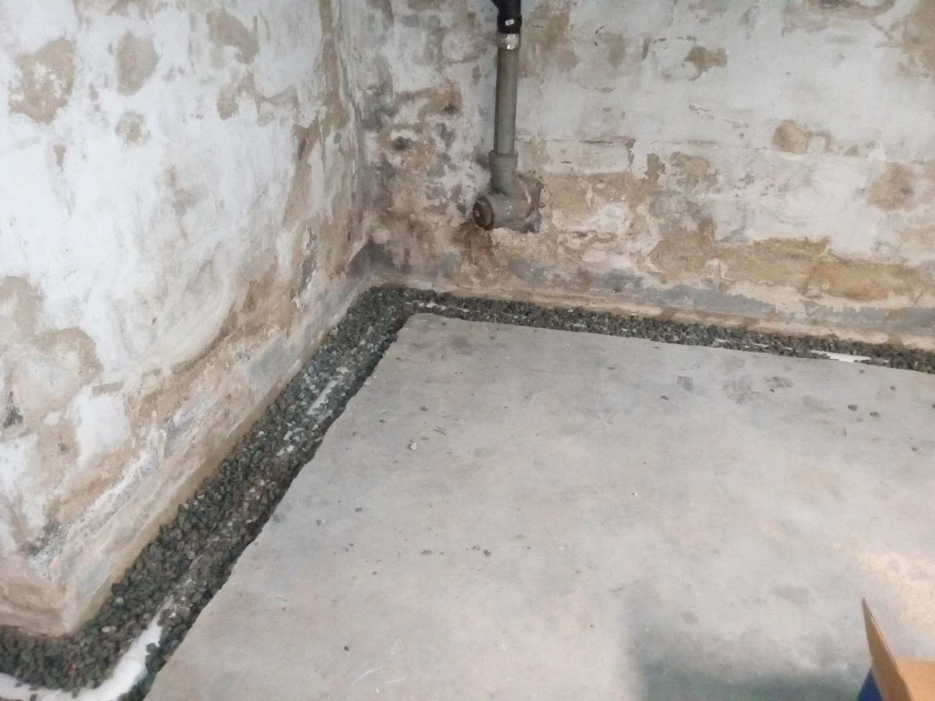 A basement corner with a gravel-filled drainage channel along the wall-to-floor joint and a pipe against the stone wall.