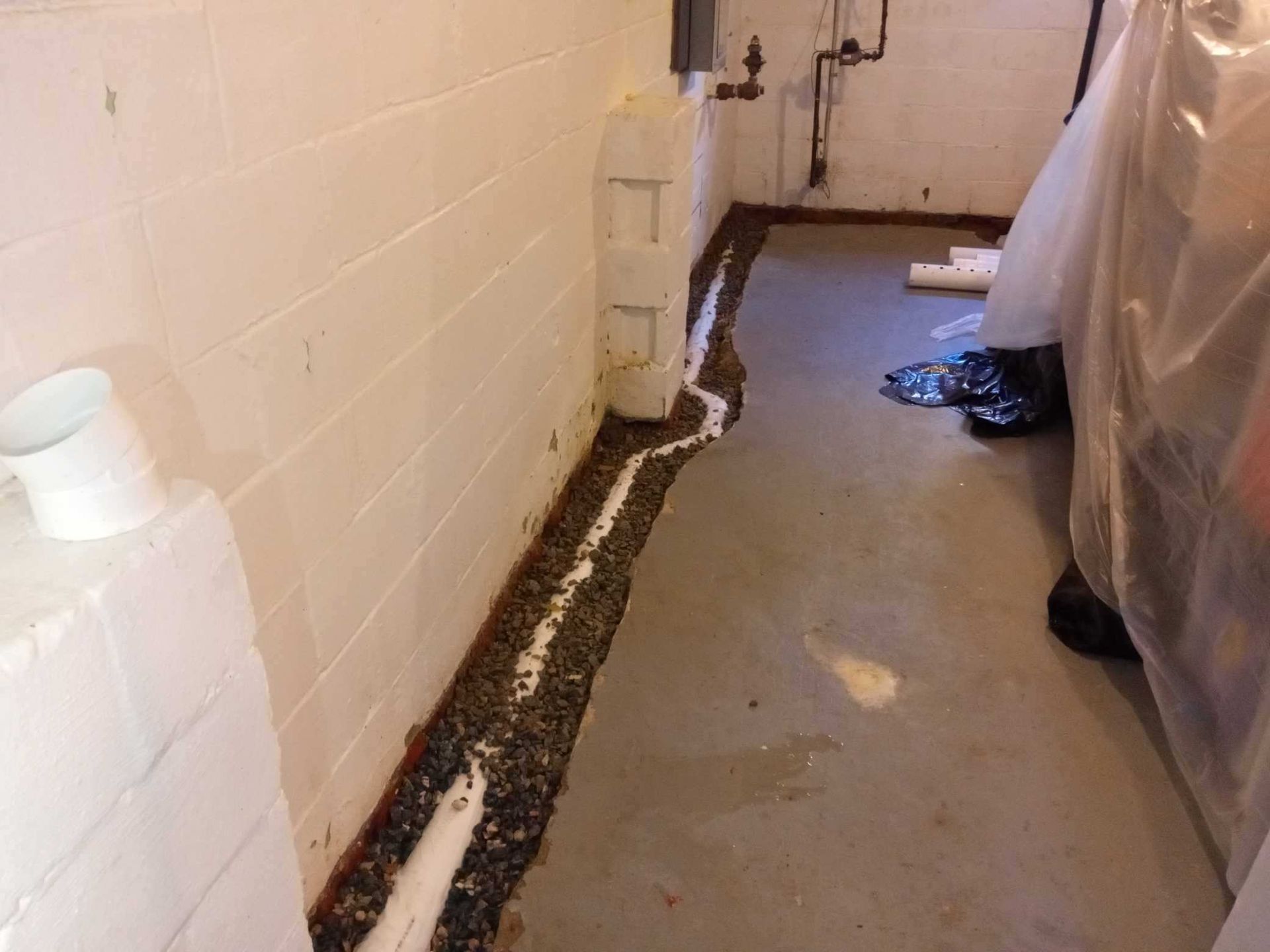 A basement perimeter with a trench cut into the concrete floor, filled with gravel and a drainage pipe near the wall.