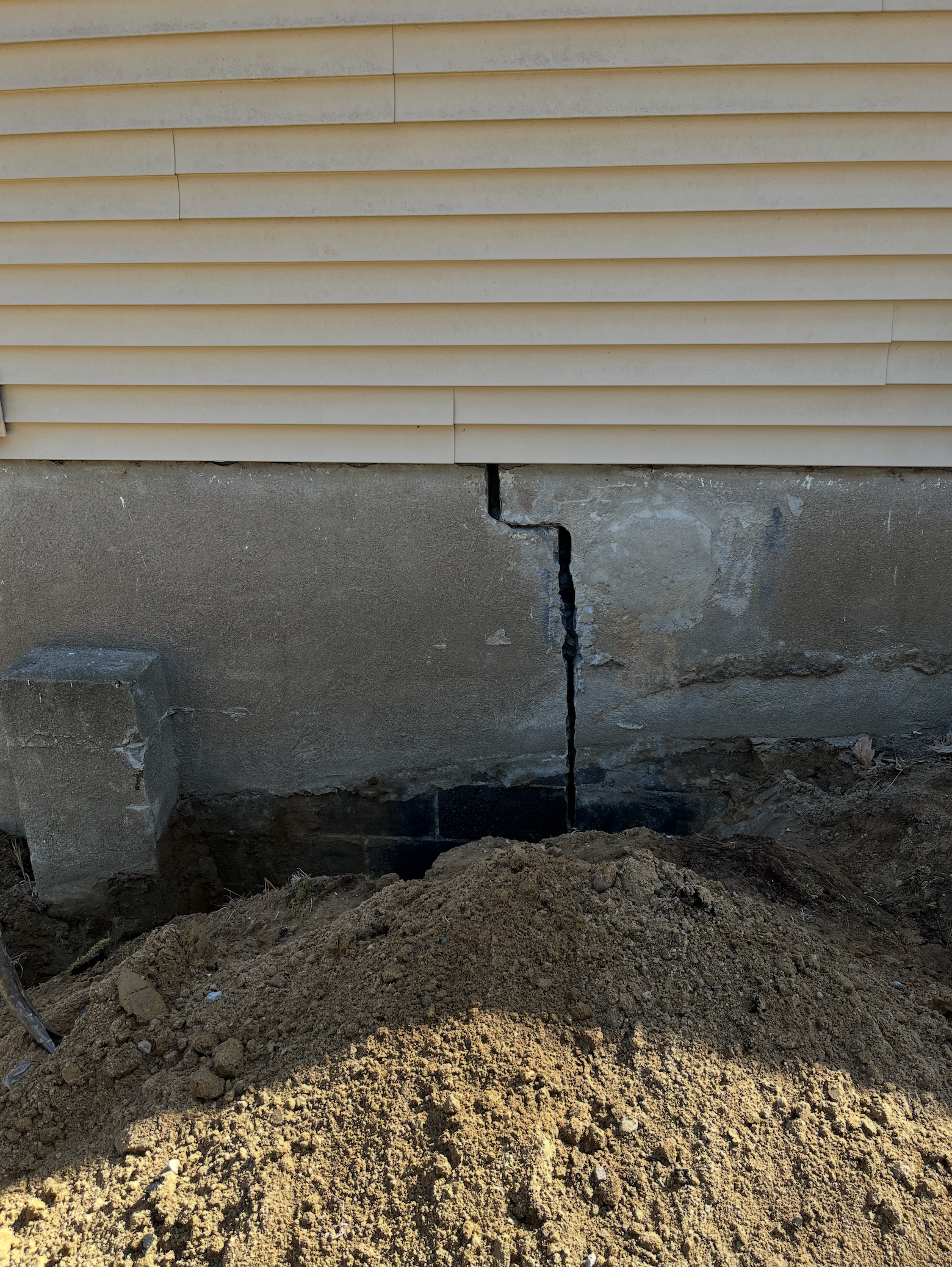 A vertical crack in a gray concrete house foundation below beige siding and a pile of dirt.