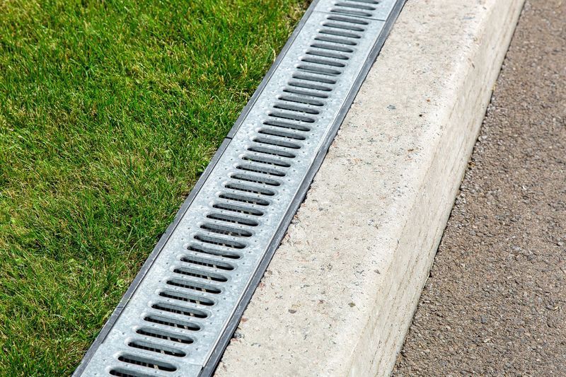 A linear metal trench drain with grates separating a patch of green grass from a concrete curb and paved walkway.