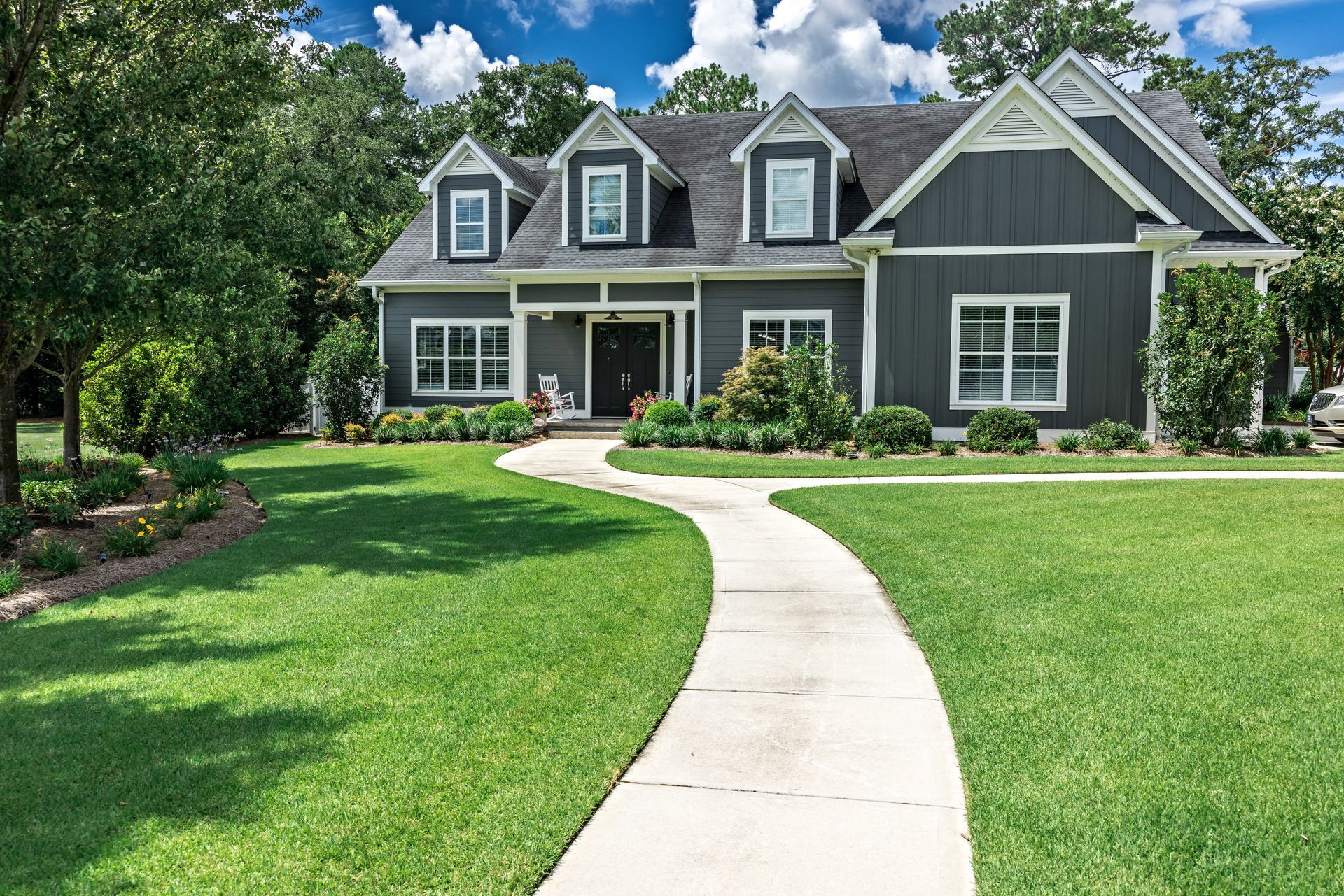 A two-story, gray house with three dormer windows and a concrete walkway through a green lawn under a sunny, blue sky.
