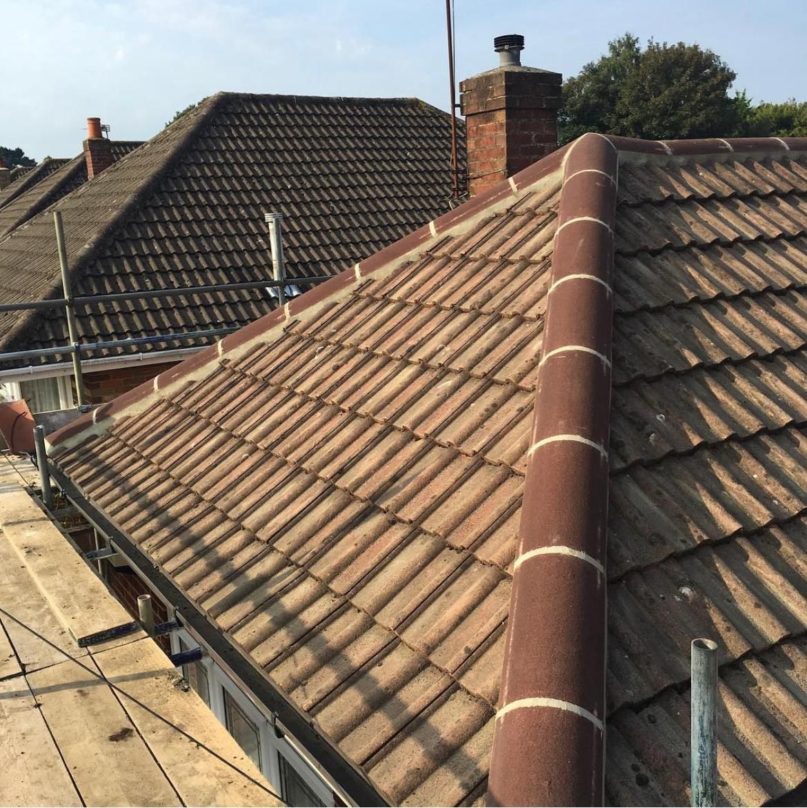 Tiled rooftops of houses with a chimney, featuring scaffolding, against a blue sky.