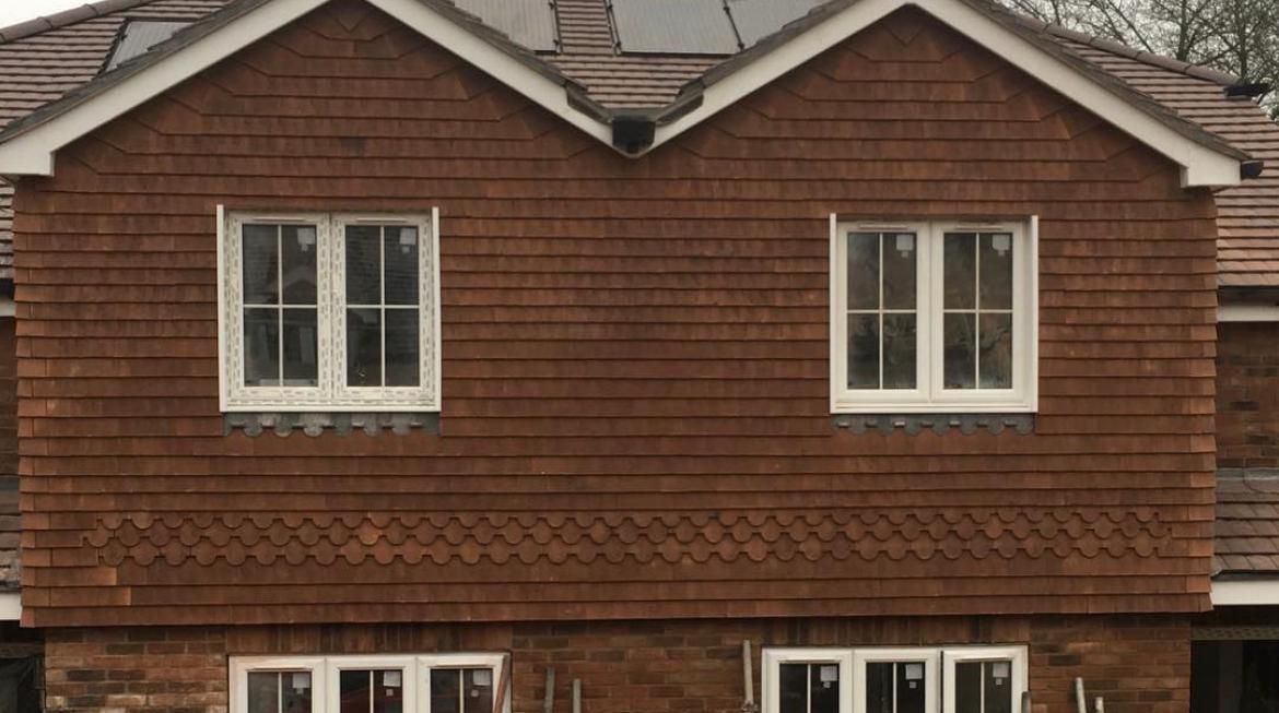 Close-up of a two-story house with brown wooden siding, white-framed windows, and a brick base.