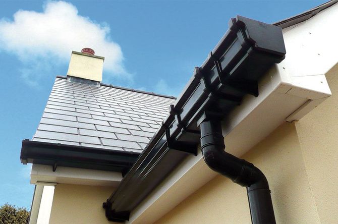 Black gutters on a light yellow house with a slate roof and clear blue sky.