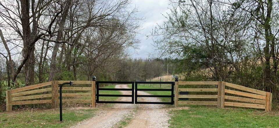 A gravel road leads to black gates in a wooden fence with trees in the background.