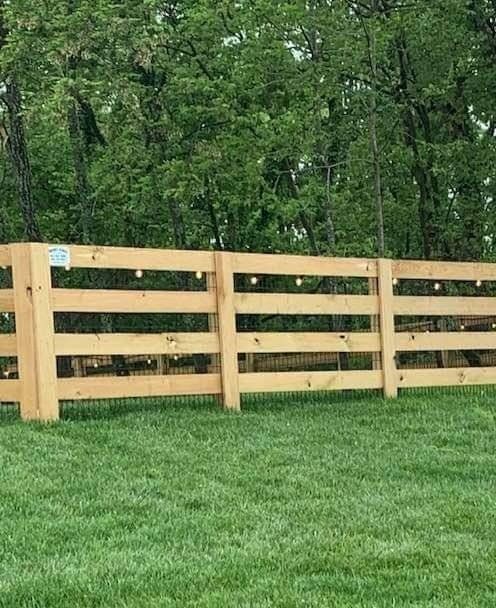 Wooden fence with string lights, on a green lawn, with a backdrop of trees.