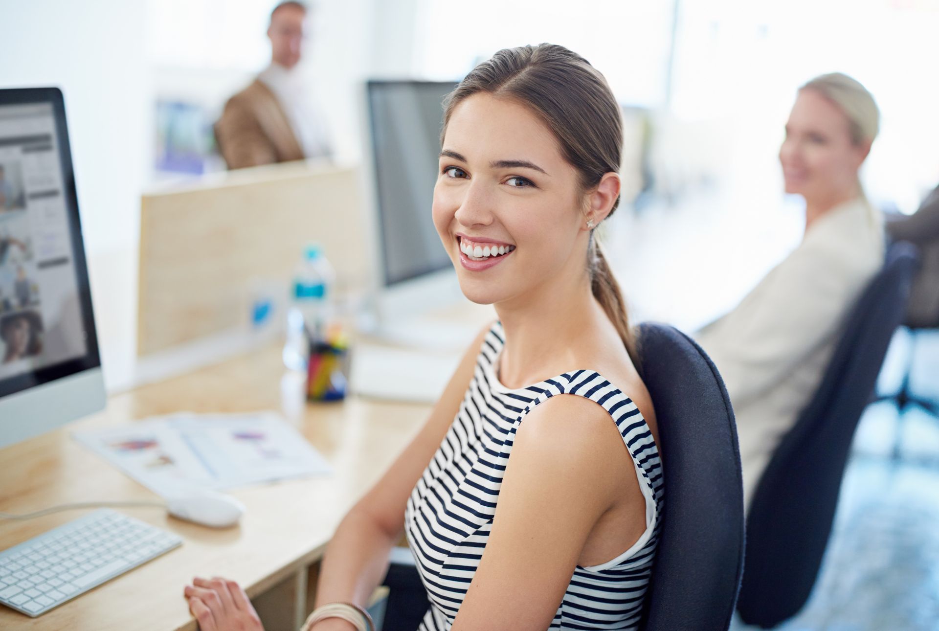 A woman is sitting at a desk in front of a computer and smiling.