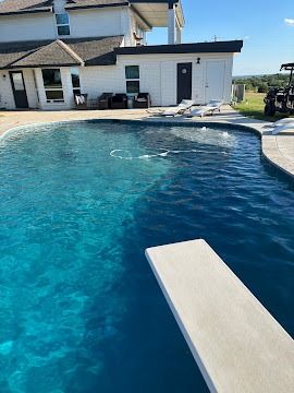 Backyard swimming pool with a diving board beside a white house on a sunny day