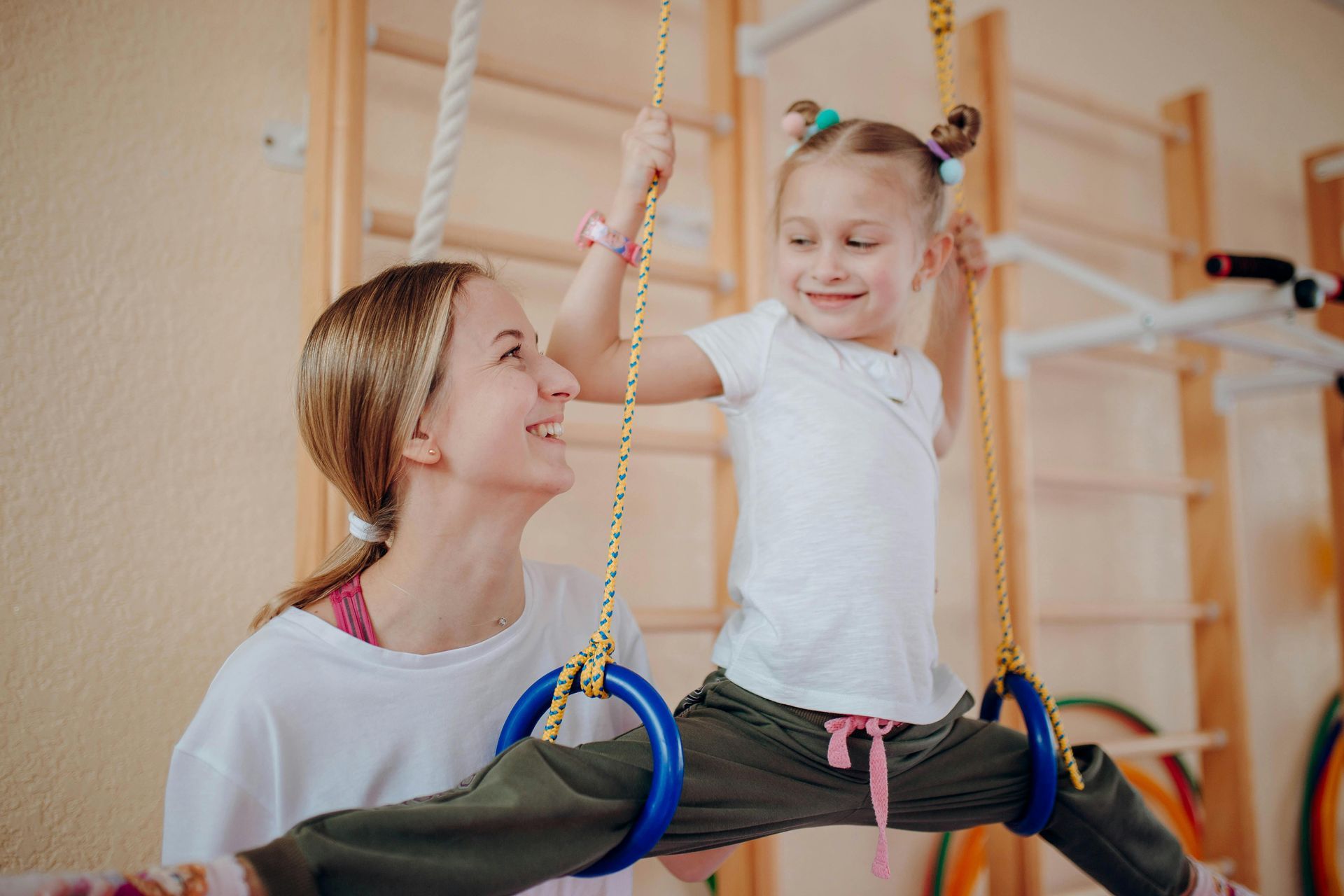 Adult and child smiling on a rope swing in a gym, holding hands, with climbing bars in the background