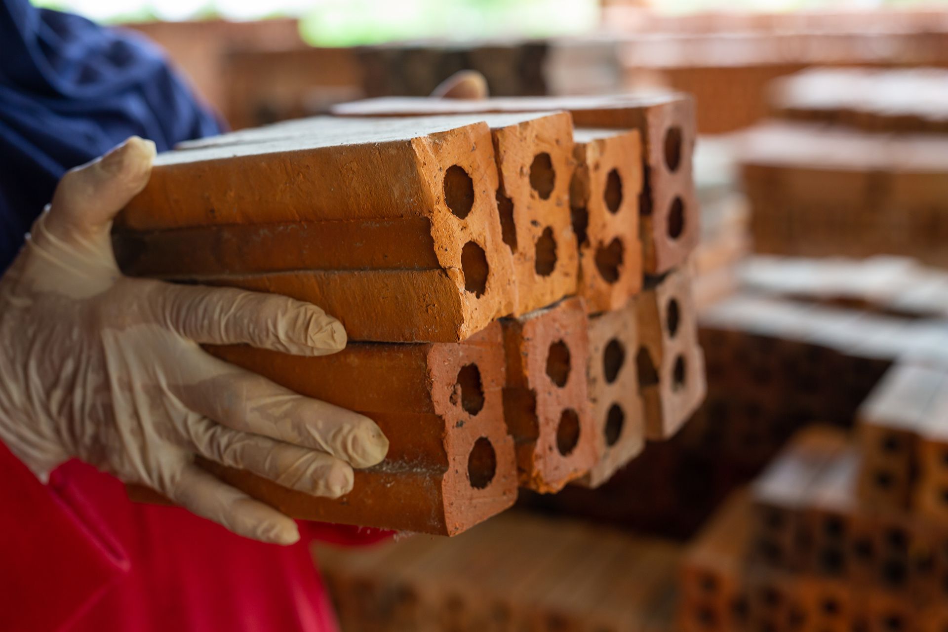 Person wearing gloves holding a stack of red bricks with holes, likely for construction.
