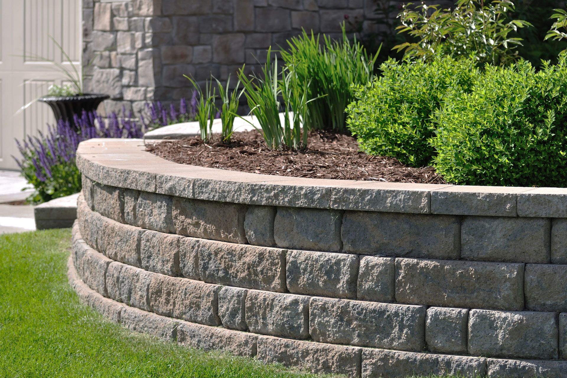 Curved stone retaining wall with a garden bed featuring plants and a lawn.