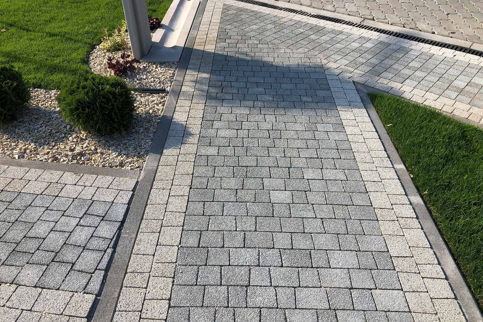 Brick walkway with gray and tan blocks bordered by green grass and plants.