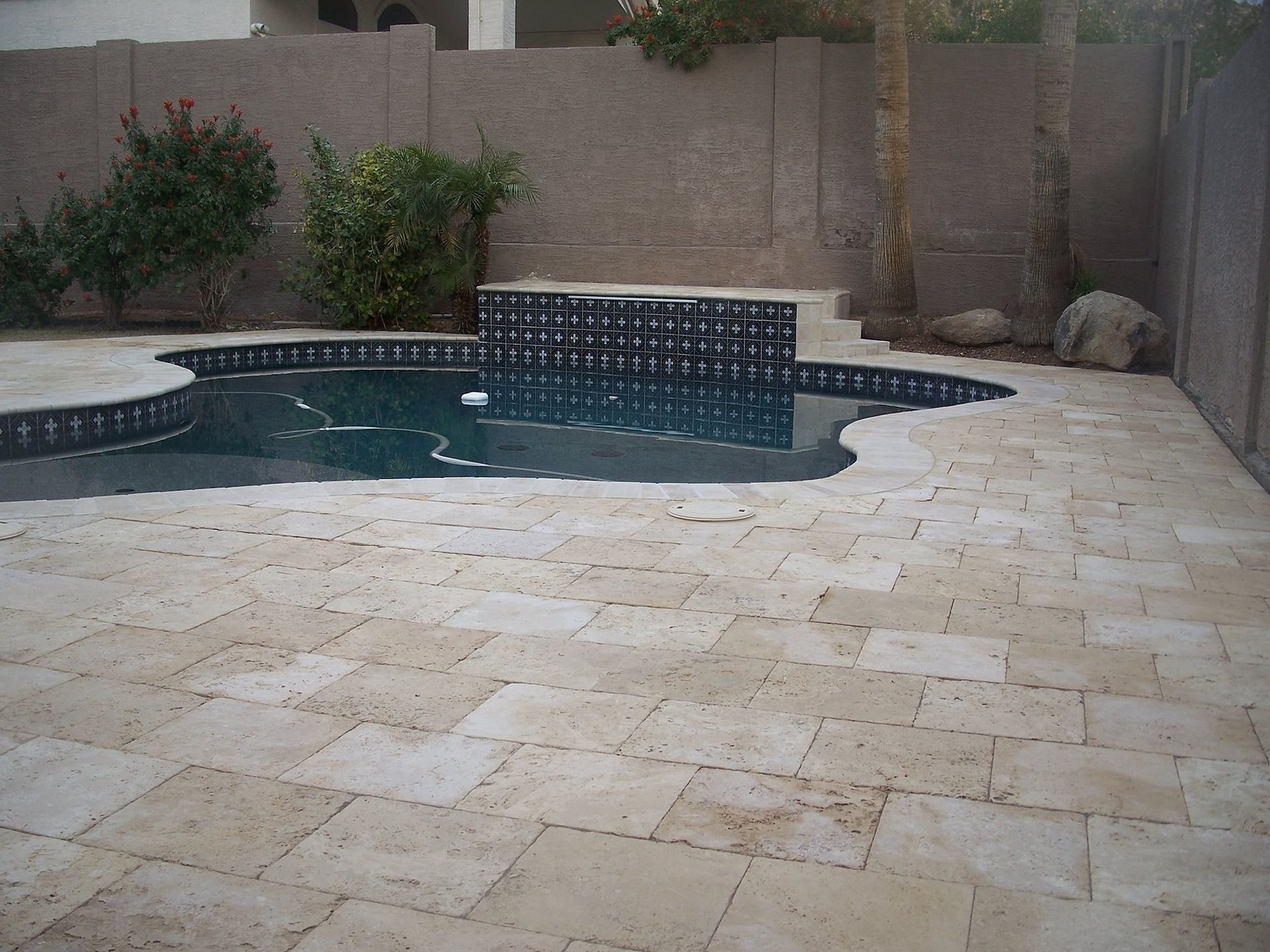 Patio with light-colored stone pavers surrounds a pool with a black tiled border and a dark water feature.
