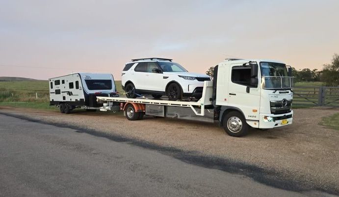 Tow Truck is Carrying a White Car on Its Flatbed and Towing a White Caravan — NCM Towing Newcastle In Broadmeadow, NSW