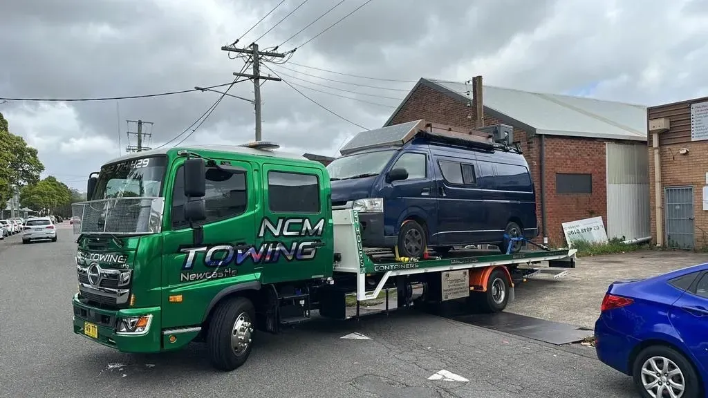 Green Towing Truck is Carrying a Blue Van Down a Street — NCM Towing Newcastle In Maitland, NSW